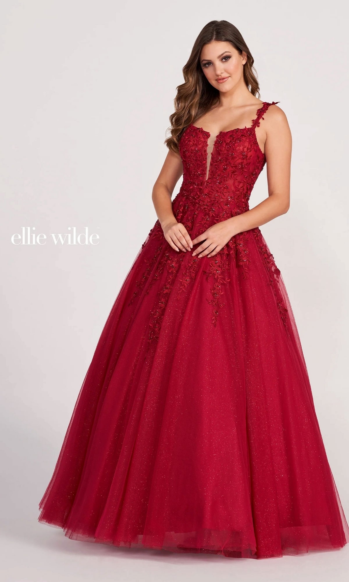 A woman wears the Ellie Wilde EW120014 long glitter prom ball gown—a sleeveless, deep red dress with floral appliqué and a full tulle skirt—standing against a plain light background. The text